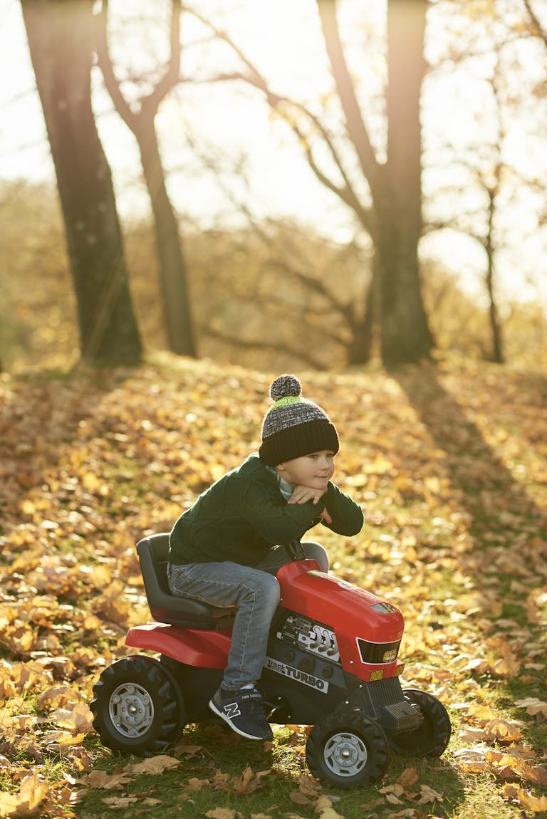 A young boy happily rides a toy tractor amidst autumn leaves in a sunny park.